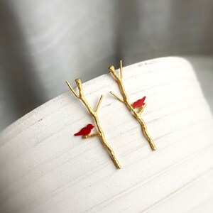 May include: A pair of gold earrings featuring a delicate branch design with a small red bird perched on each branch.