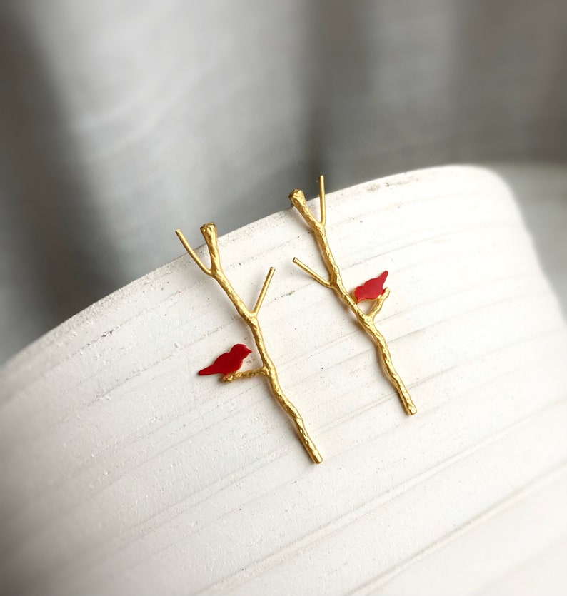 May include: A pair of gold earrings featuring a delicate branch design with a small red bird perched on each branch.