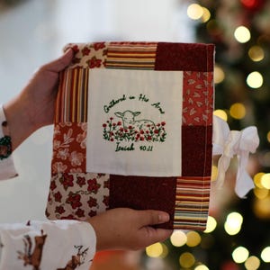 May include: A patchwork book cover with a central embroidered design featuring a lamb and the text "Gathered in His Arms, Isaiah 40:11." The cover combines floral and striped patterns in red, cream, and burgundy. A white ribbon is attached.