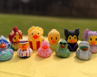 Könnte beinhalten: Eine Sammlung von zehn kleinen Quietscheentchen, die jeweils mit bunten Strasssteinen verziert sind. Die Enten zeigen verschiedene Designs, darunter einen Popcorn-Eimer, eine schwarze Katze, ein rosa Punktemuster und weitere Kostüme mit Kristallen.