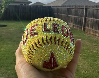 Puede incluir: Una pelota de sóftbol amarilla completamente cubierta de pedrería iridiscente. La pelota presenta el texto DE LEON en pedrería roja en la parte superior y una letra J en pedrería roja en la parte inferior, con detalles de costura.