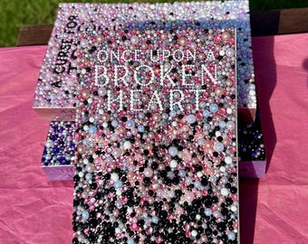 Könnte beinhalten: Zwei dekorative Boxen mit dem Text "ONCE UPON A BROKEN HEART" auf der Vorderseite. Die Boxen sind mit einem Mosaik aus rosa, weißen und schwarzen Perlen bedeckt. Die Boxen stehen auf einer rosa Oberfläche.