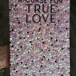 Puede incluir: Una obra de arte rectangular con el texto "A Curse For True Love" en letras negras. La superficie est&aacute; cubierta con una mezcla de cuentas y perlas blancas, rosas e iridiscentes, creando un efecto texturizado y brillante. La est&eacute;tica general es rom&aacute;ntica y decorativa.