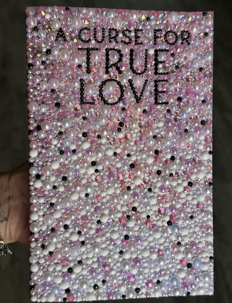 Puede incluir: Una obra de arte rectangular con el texto "A Curse For True Love" en letras negras. La superficie est&aacute; cubierta con una mezcla de cuentas y perlas blancas, rosas e iridiscentes, creando un efecto texturizado y brillante. La est&eacute;tica general es rom&aacute;ntica y decorativa.