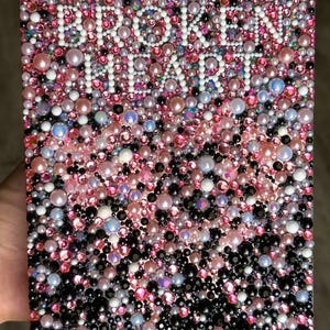 Puede incluir: Una obra de arte rectangular cubierta por un mosaico de cuentas rosas, blancas, negras e iridiscentes. Las palabras "ONCE UPON A BROKEN HEART" est&aacute;n escritas con cuentas blancas en el centro de la pieza.