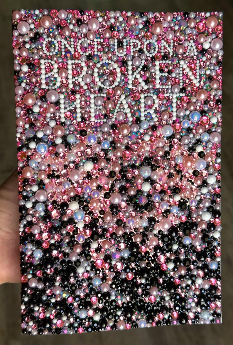 Puede incluir: Una obra de arte rectangular cubierta por un mosaico de cuentas rosas, blancas, negras e iridiscentes. Las palabras "ONCE UPON A BROKEN HEART" est&aacute;n escritas con cuentas blancas en el centro de la pieza.