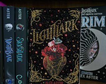 Könnte beinhalten: Ein Hardcover-Buch mit dem Titel "Nightmark" von Alex Aster, mit einem schwarzen Einband, verziert mit goldenen und roten Perlen. Das Cover zeigt ein florales Design und goldene Schrift. Weitere Bücher sind im Regal sichtbar.