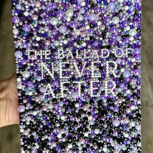 Puede incluir: Una obra de arte rectangular cubierta por un mosaico de cuentas moradas, blancas y negras. El texto "THE BALLAD OF NEVER AFTER" est&aacute; escrito con cuentas blancas. La obra de arte est&aacute; sostenida por una mano.