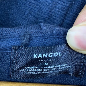 Op de afbeelding: Een donkerblauw kledingstuk met capuchon en een zwart rechthoekig stoffen label dat aan de binnenkant van de halslijn is gestikt. Het label bevat de witte tekst KANGOL, roshell, M en aanvullende handelsmerkinformatie in een kleiner lettertype.