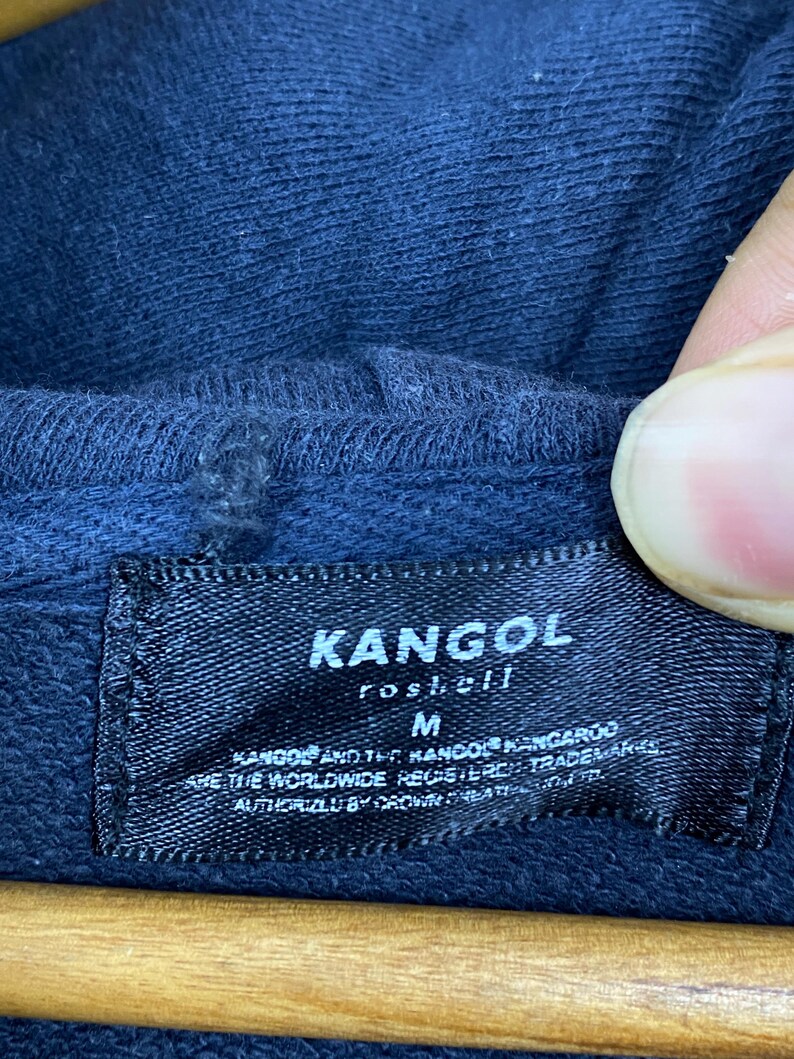 Op de afbeelding: Een donkerblauw kledingstuk met capuchon en een zwart rechthoekig stoffen label dat aan de binnenkant van de halslijn is gestikt. Het label bevat de witte tekst KANGOL, roshell, M en aanvullende handelsmerkinformatie in een kleiner lettertype.
