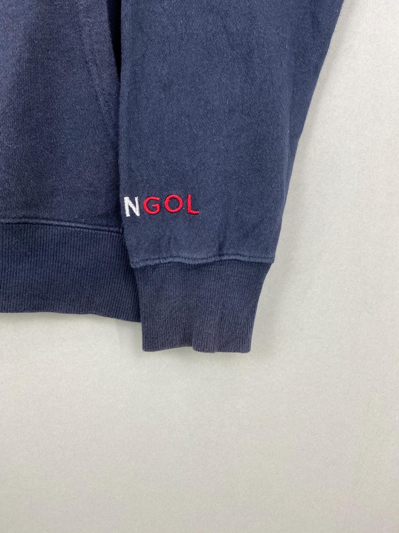 Op de afbeelding: Een marineblauw sweatshirt met lange mouwen, voorzien van een geribbelde boord en zoom. Het woord NGOL is geborduurd in wit en rood garen op de mouw bij de boord. De stof heeft een zichtbare textuur en het kledingstuk heeft een klassieke ronde hals.