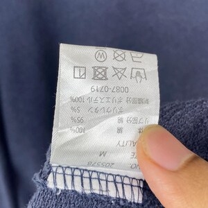Op de afbeelding: Een wit rechthoekig waslabel bevestigd aan een donkerblauwe stof. Het label bevat zwarte tekst, waaronder maat M, productnummer 205578 en diverse wassymbolen aan de bovenzijde. De stof heeft een textuur die lijkt op gebreid materiaal.