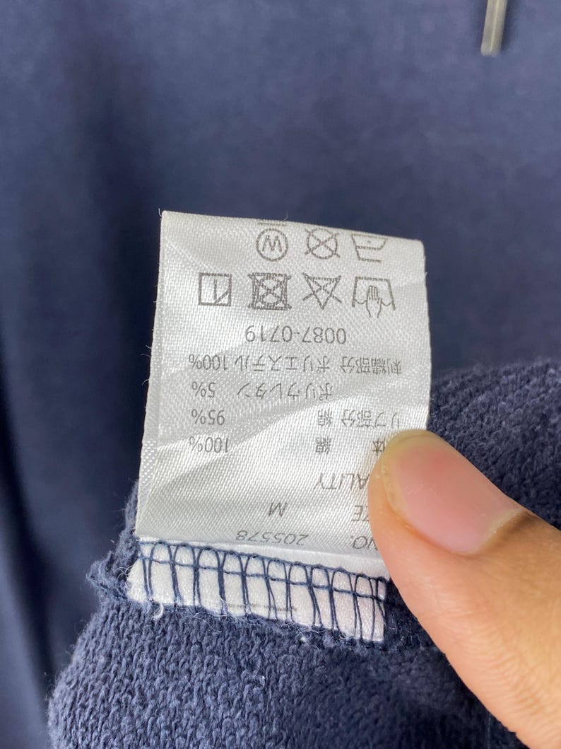 Op de afbeelding: Een wit rechthoekig waslabel bevestigd aan een donkerblauwe stof. Het label bevat zwarte tekst, waaronder maat M, productnummer 205578 en diverse wassymbolen aan de bovenzijde. De stof heeft een textuur die lijkt op gebreid materiaal.