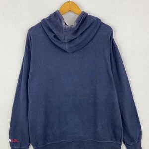 Op de afbeelding: Een marineblauwe hoodie met lange mouwen en een capuchon. De stof heeft een vervaagde uitstraling. De tekst "NGOL" is in het rood gedrukt op het onderste gedeelte van de rechtermouw. Het kledingstuk hangt aan een houten kledinghanger.