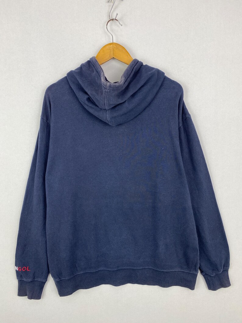 Op de afbeelding: Een marineblauwe hoodie met lange mouwen en een capuchon. De stof heeft een vervaagde uitstraling. De tekst "NGOL" is in het rood gedrukt op het onderste gedeelte van de rechtermouw. Het kledingstuk hangt aan een houten kledinghanger.