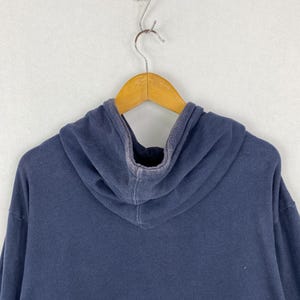 Op de afbeelding: Een effen marineblauwe hoodie met lange mouwen en een capuchon, voorzien van een licht vervaagde, versleten textuur langs de naden en randen. Het kledingstuk is gemaakt van zachte stof en hangt aan een houten kledinghanger.