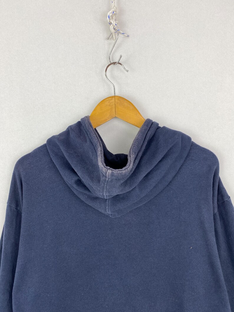 Op de afbeelding: Een effen marineblauwe hoodie met lange mouwen en een capuchon, voorzien van een licht vervaagde, versleten textuur langs de naden en randen. Het kledingstuk is gemaakt van zachte stof en hangt aan een houten kledinghanger.