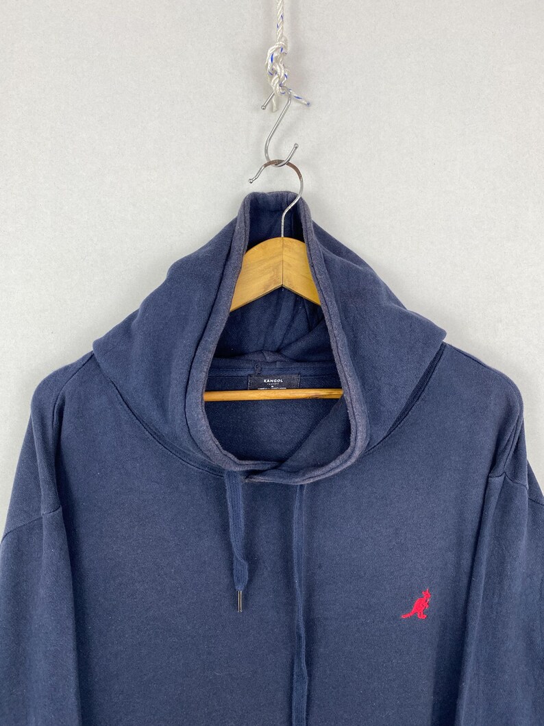 Op de afbeelding: Een marineblauw sweatshirt met capuchon, een hoge kraag en een trekkoord. Het kledingstuk is voorzien van een klein, geborduurd rood kangoeroelogo aan de rechteronderkant en een Kangol-merklabel aan de binnenkant van de kraag.
