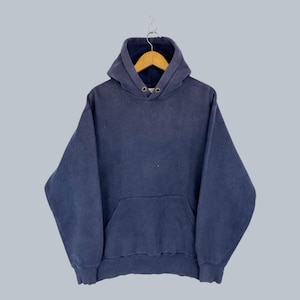 Könnte beinhalten: Ein marineblauer Kapuzenpullover mit Kängurutasche und Kapuze mit Kordelzug. Das Sweatshirt hängt an einem Holzbügel vor einem hellgrauen Hintergrund. Der Hoodie ist aus einem weichen, bequemen Material gefertigt.