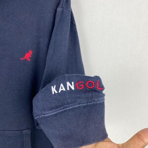 Op de afbeelding: Een marineblauw sweatshirt met lange mouwen, voorzien van een rood geborduurd kangoeroelogo op de borst en de tekst KANGOL in wit en rood geborduurd op de geribbelde boord van de mouw. Het kledingstuk heeft een klassieke ronde halslijn.