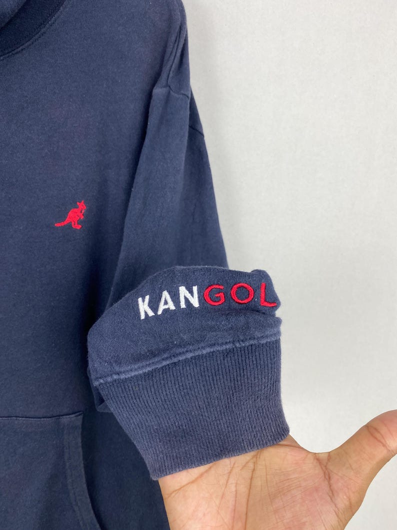 Op de afbeelding: Een marineblauw sweatshirt met lange mouwen, voorzien van een rood geborduurd kangoeroelogo op de borst en de tekst KANGOL in wit en rood geborduurd op de geribbelde boord van de mouw. Het kledingstuk heeft een klassieke ronde halslijn.