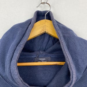 Op de afbeelding: Een marineblauwe hoodie met een donker stoffen label aan de binnenkant van de halslijn met de witte tekst KANGOL en de maat M eronder gedrukt. Het kledingstuk heeft een dikke capuchon met een bijpassend trekkoord en een zichtbare naad.