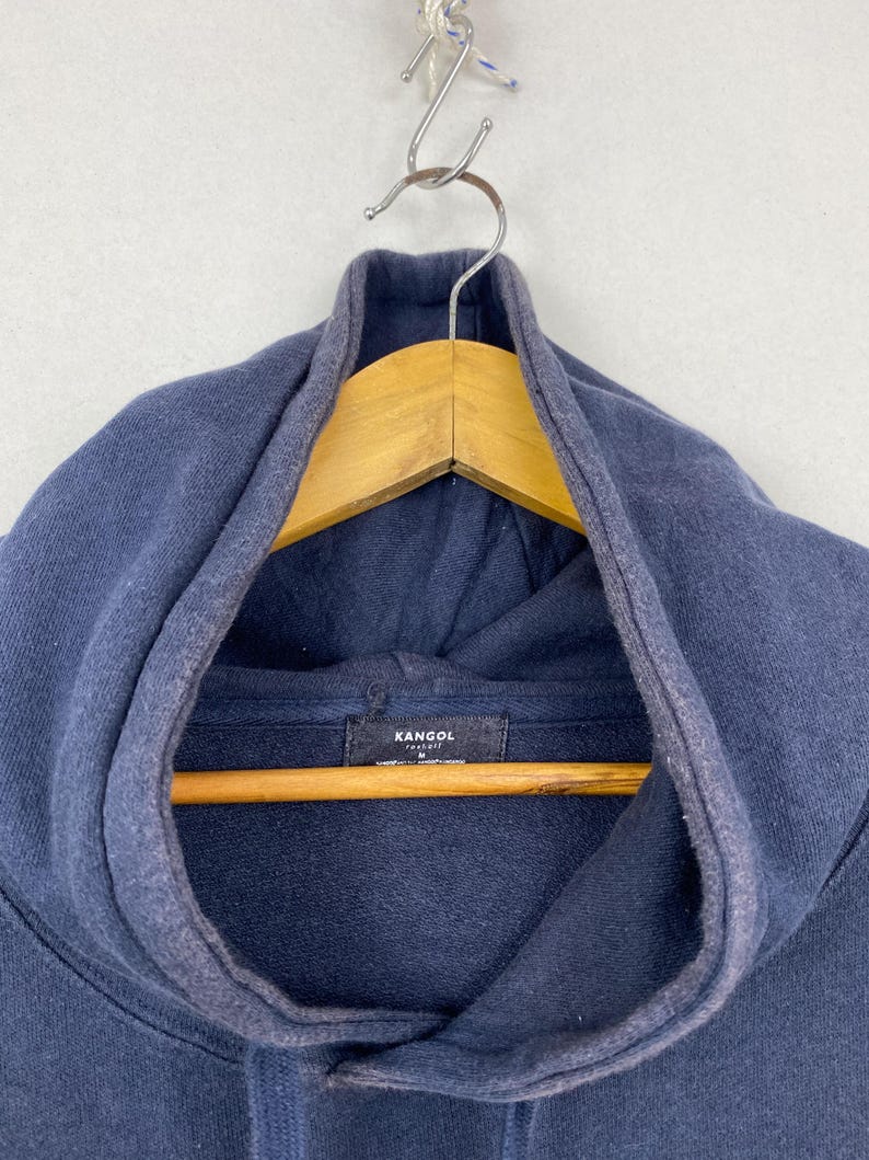 Op de afbeelding: Een marineblauwe hoodie met een donker stoffen label aan de binnenkant van de halslijn met de witte tekst KANGOL en de maat M eronder gedrukt. Het kledingstuk heeft een dikke capuchon met een bijpassend trekkoord en een zichtbare naad.