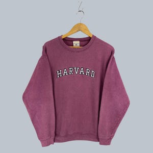 May include: A long-sleeve crewneck sweatshirt in a faded plum color. The front features the word HARVARD embroidered in white capital letters with a black outline in an arched design. The garment includes a ribbed collar, cuffs, and hem.