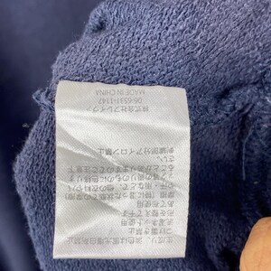 Op de afbeelding: Een wit rechthoekig waslabel genaaid op een donkerblauwe stof. Het label bevat zwarte Japanse tekst met was- en onderhoudsinstructies, evenals de tekst 'MADE IN CHINA' en het nummer '06-6531-1147'.