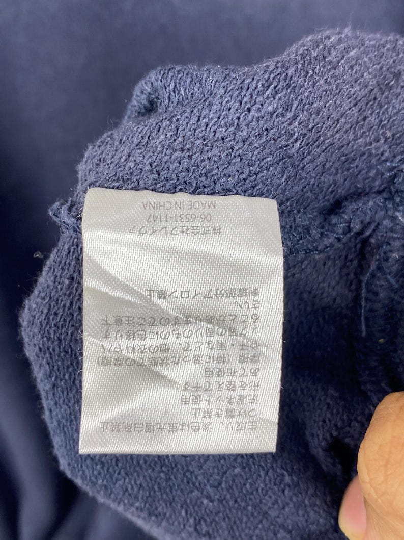 Op de afbeelding: Een wit rechthoekig waslabel genaaid op een donkerblauwe stof. Het label bevat zwarte Japanse tekst met was- en onderhoudsinstructies, evenals de tekst 'MADE IN CHINA' en het nummer '06-6531-1147'.