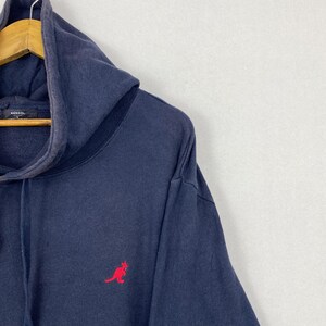 Op de afbeelding: Een marineblauwe hoodie met een rood geborduurd kangoeroelogo op de borst. Het kledingstuk heeft een capuchon met trekkoord en een Kangol-merklabel zichtbaar aan de binnenkant van de halslijn, met een vervaagde, gedragen textuur.