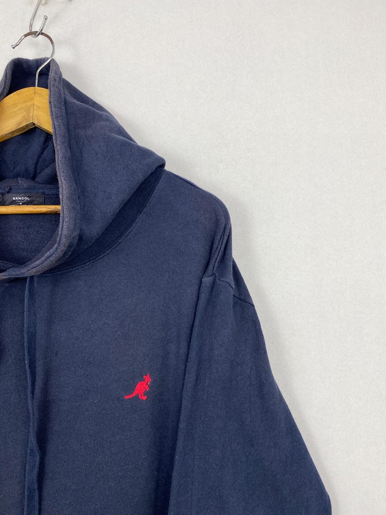 Op de afbeelding: Een marineblauwe hoodie met een rood geborduurd kangoeroelogo op de borst. Het kledingstuk heeft een capuchon met trekkoord en een Kangol-merklabel zichtbaar aan de binnenkant van de halslijn, met een vervaagde, gedragen textuur.