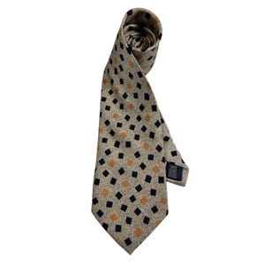Puede incluir: Una corbata con fondo beige texturizado y un patrón repetitivo de pequeños rombos de color azul marino oscuro y naranja quemado. La corbata está enrollada en la parte superior e incluye una pequeña etiqueta de tela rectangular oscura en el reverso.
