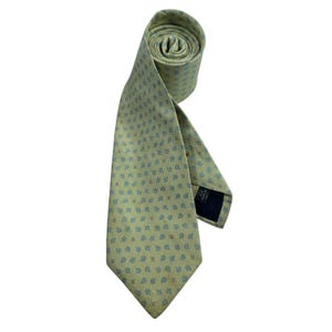 May include: A light green necktie featuring a repeating pattern of small blue floral motifs. The tie is rolled at the top and includes a dark label on the back that reads Made in Italy. The fabric has a subtle texture throughout the entire length.
