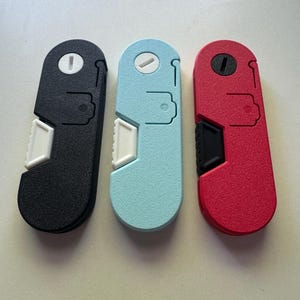 May include: Three multi-tool gadgets in black, light blue, and red. Each tool has a rounded top with a circular feature, a white rectangular button, and a black or red angled section. The tools are designed with a textured surface.