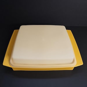 May include: A vintage yellow and cream-colored butter dish. The dish has a square shape with a yellow base and a cream-colored lid. The lid sits on top of the base, and the dish is made of plastic.