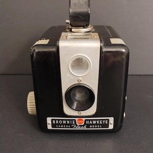May include: Vintage Kodak Brownie Hawkeye Flash camera. The black camera has a silver faceplate with a lens and viewfinder. The camera has a handle on top and the text "Brownie Hawkeye Camera Flash Model" on the front.