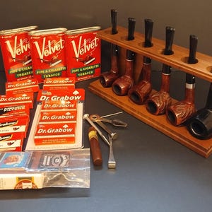 May include: A collection of six tobacco pipes displayed on a wooden rack, accompanied by three red Velvet tobacco tins, several boxes of Dr. Grabow pipe filters, a metal pipe tool, a small cigar, and a pack of matches in a plastic bag.