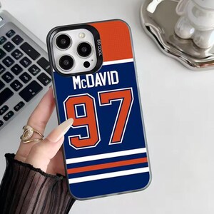 May include: A phone case featuring a blue and orange jersey design with the name "McDavid" and the number "97". The case has a black border and the words "SO COOL" are printed on the side.
