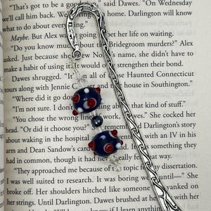 May include: A silver metal bookmark with a decorative design and a beaded charm. The charm features dark blue and red glass beads. The bookmark is resting on the pages of a book.