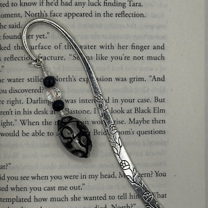 May include: A silver metal bookmark with a floral design and a beaded charm. The charm features black and clear beads and a black and gray oval-shaped bead. The bookmark is resting on the pages of a book.