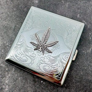May include: A rectangular silver-toned metal cigarette case featuring an intricate engraved scrollwork pattern across the surface and a prominent, raised three-dimensional cannabis leaf emblem centered on the front cover of the container.