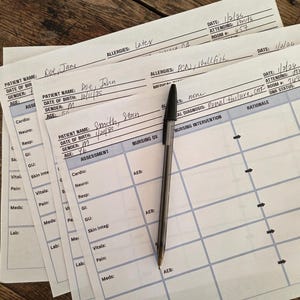 May include: A stack of patient assessment forms with handwritten information and a black pen on a wooden surface. The forms include fields for patient name, date of birth, and medical information. The pen is positioned vertically.