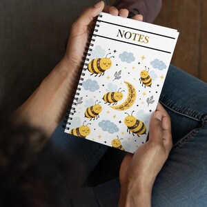 Puede incluir: Un cuaderno con espiral con una cubierta blanca que presenta un patrón repetido de abejas de dibujos animados, nubes y una luna creciente. La palabra "NOTES" está impresa en oro en la parte superior. Las abejas son amarillas y negras.