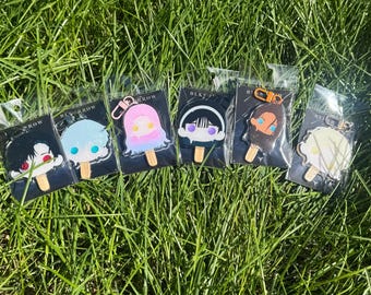 May include: Six acrylic keychains shaped like popsicles, each featuring a unique character head design with a small wooden stick base. The keychains are individually packaged in clear plastic sleeves and attached to black backing cards.