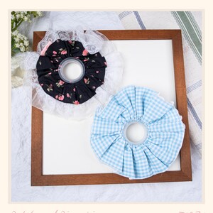 May include: Two handmade pincushions. One is black with a floral pattern and white lace trim. The other is light blue gingham with white lace trim. The pincushions are on a white background inside a wooden frame. The text "Petals and Pincushions" is at the bottom.