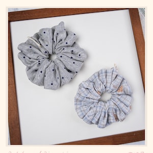 May include: Two fabric scrunchies. One is gray with navy blue polka dots. The other is blue and white plaid with small brown bear graphics. The scrunchies are on a white background inside a wooden frame. Text at the bottom reads "Petals and Pincushions - TAO21".