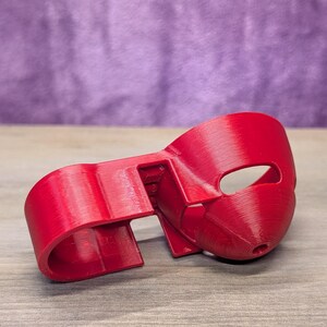 Op de afbeelding: Een felrood, 3D-geprint masker met een oogopening en een gebogen ontwerp. Het masker ligt op een houten oppervlak met een paarse achtergrond.