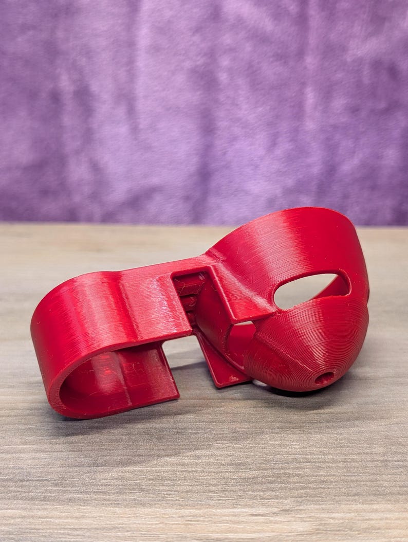 Op de afbeelding: Een felrood, 3D-geprint masker met een oogopening en een gebogen ontwerp. Het masker ligt op een houten oppervlak met een paarse achtergrond.