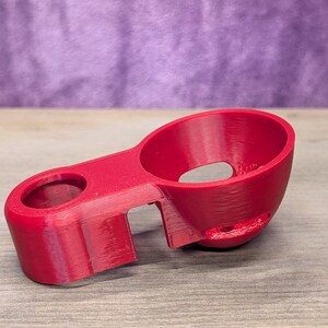 Op de afbeelding: Een helder rood 3D-geprint object met twee cirkelvormige openingen en een verbindingsbrug. Het object heeft een gebogen ontwerp en staat op een houten oppervlak, met een paarse achtergrond.